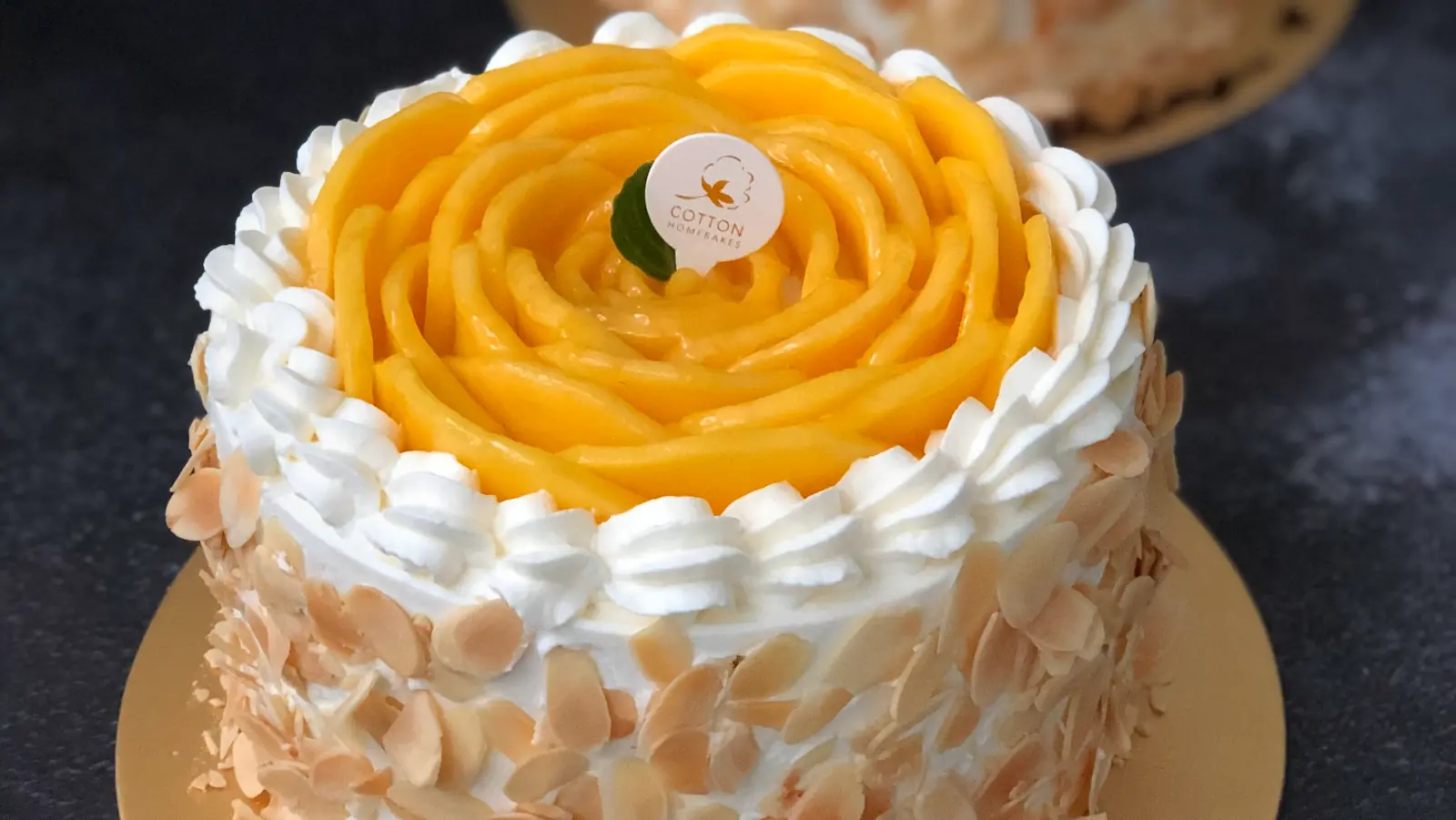Mango cake topped with a rose of fresh mango slices, piped cream and toasted almond flakes